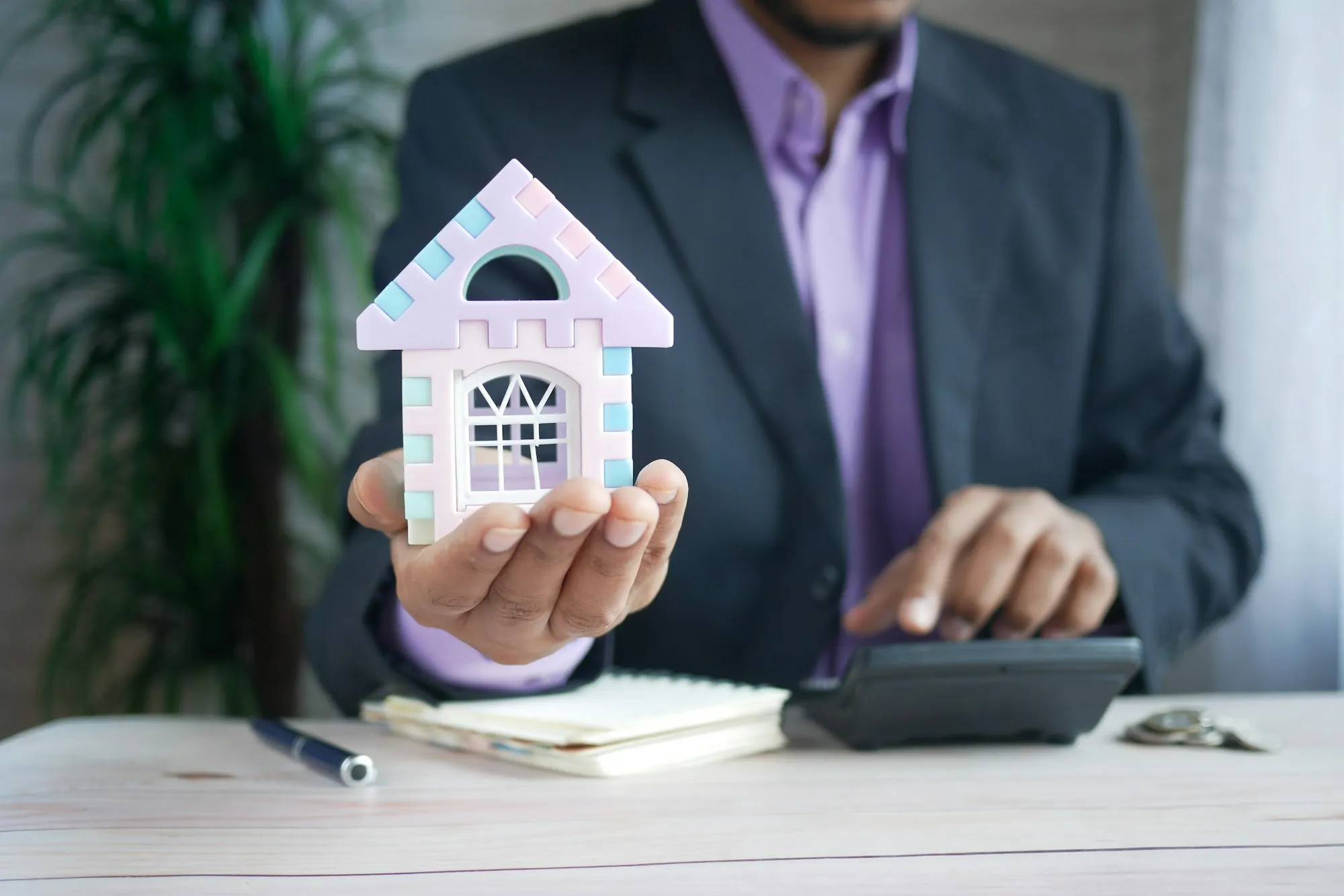 Person holding a model house beside a calculator and notebook